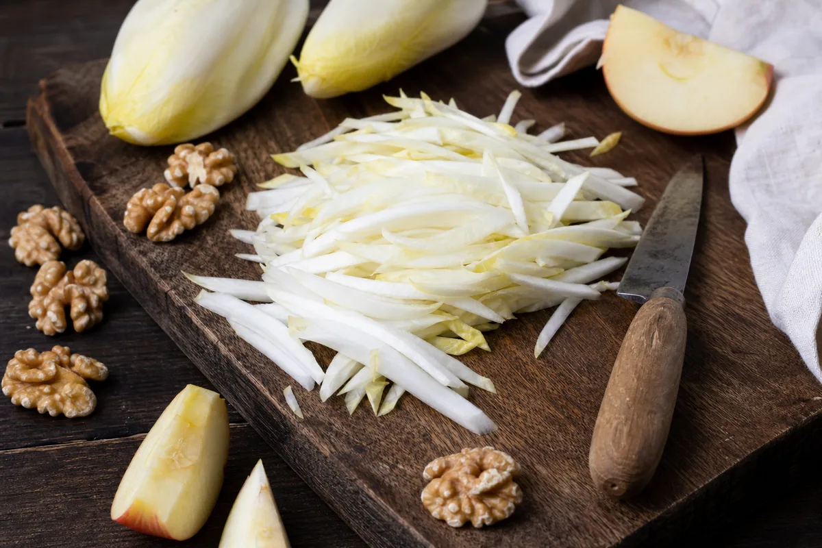 Chicons crus tranchés en lamelles sur une planche en bois, à côté de noix et de pommes